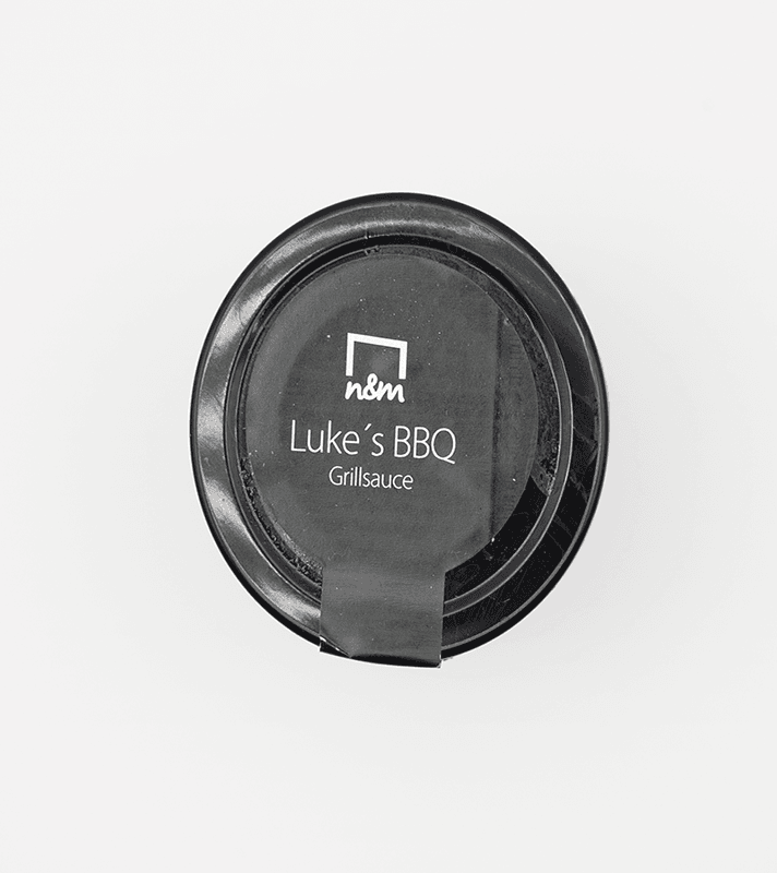 Schwarze runde Grillsauce-Box mit weißem Etikett, auf dem "nom Luke's BBQ Grillsauce" steht, mit schwarzem Deckel und abgedecktem Verschluss.