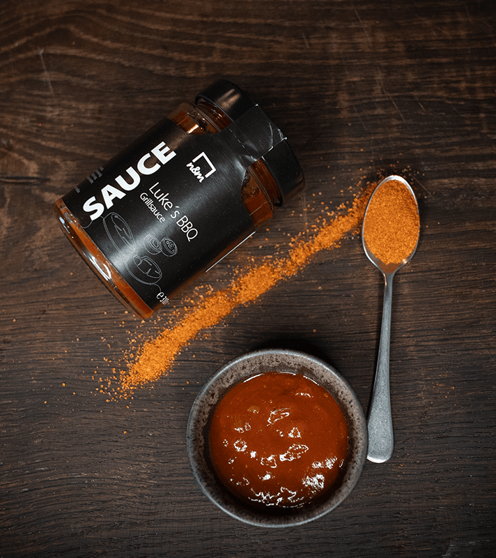 Glas mit BBQ-Sauce, daneben ein Löffel mit Paprikapulver, und ein Gewürzglas mit BBQ-Sauce auf dunklem Holzuntergrund.