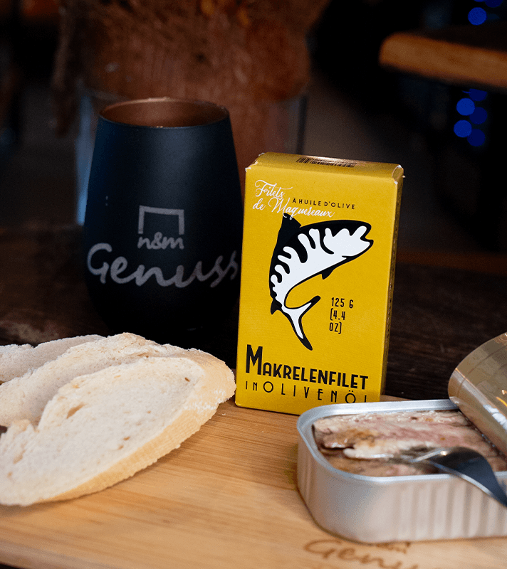 Schwarzer Becher mit weißem Schriftzug "Genuss", offene Dose Makrelenfilet in Olivenöl, drei Brotscheiben auf Holzbrett, und eine halb geöffnete Fischdose mit Löffel.