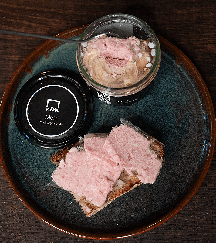 Glas mit rohem Fleisch, auf einem Teller mit Brotscheiben, daneben ein Deckel mit dem Aufdruck "Mett im Geleemantel".