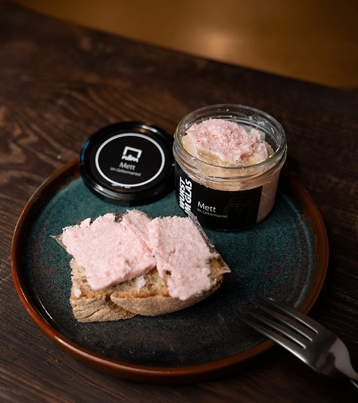 Glas mit rohem Fleisch, auf einem Teller mit Brotscheiben, daneben ein Deckel mit dem Aufdruck "Mett im Geleemantel".