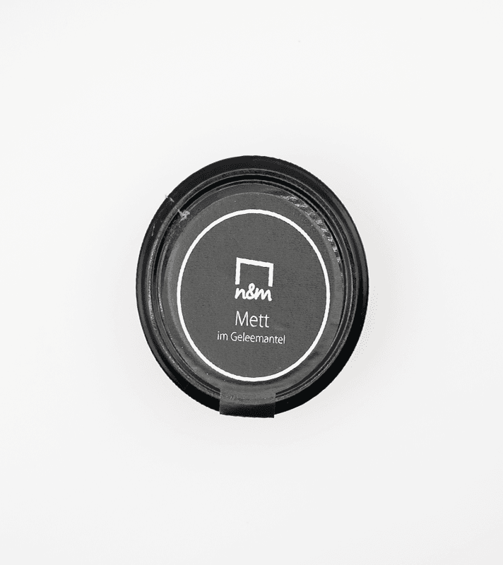 Runder schwarzer Deckel mit weißem Label, das "nsm Mett im Geleemantel" beschreibt, auf weißem Hintergrund.