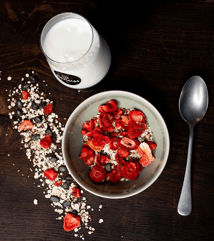 Ein Teller mit Müsli, Erdbeeren und Samen, daneben eine Glas Milch und ein Löffel auf dunklem Holz.