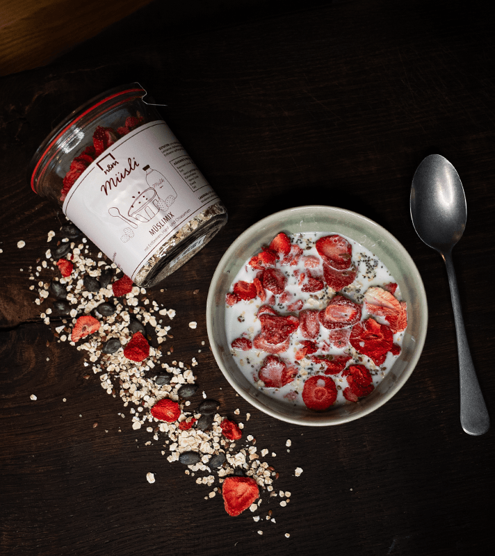 Ein Teller mit Müsli, Erdbeeren und Samen, daneben eine Glas Milch und ein Löffel auf dunklem Holz.
