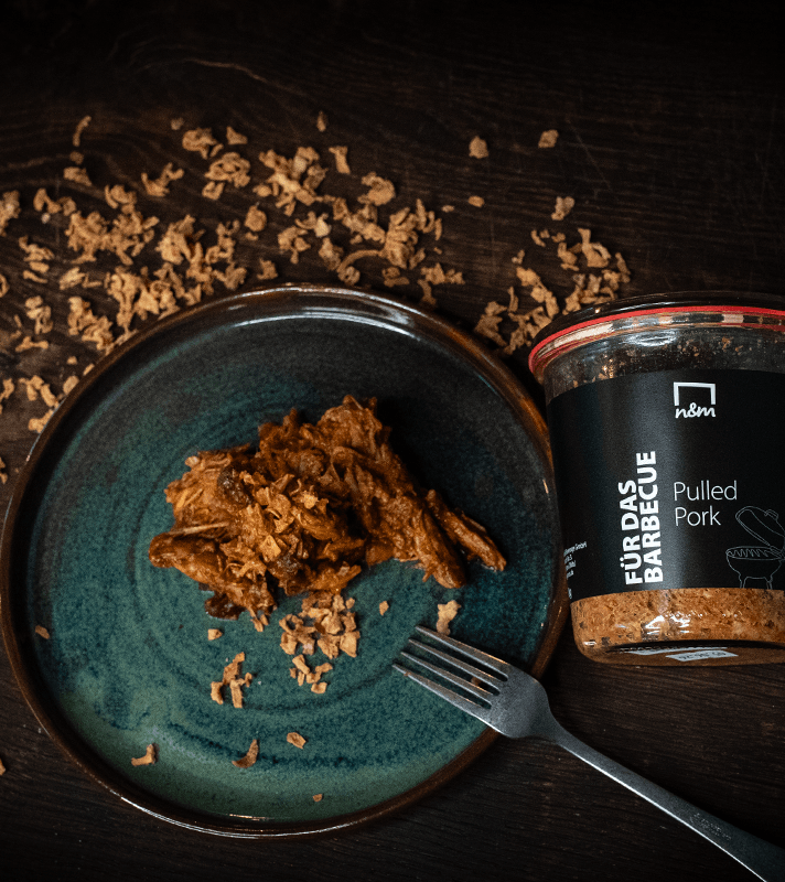 Kleines, grüner Teller mit zerkleinertem Pulled Pork in Barbecue-Sauce, daneben ein Glas mit schwarzem Etikett, auf dunklem Holzuntergrund mit verstreuten Bröseln.