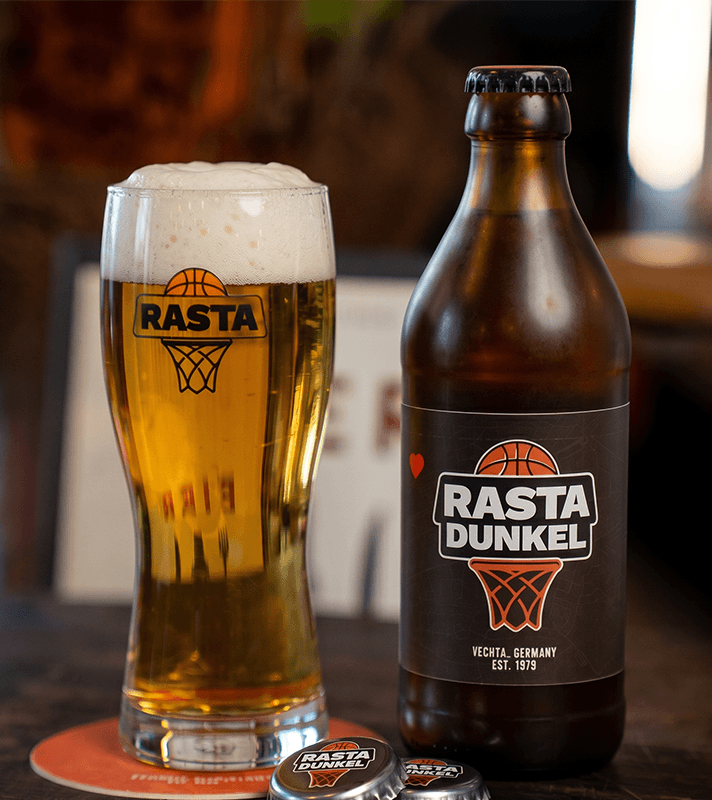 Eine braune Bierflasche mit Rasta Dunkel-Logo, daneben ein gefülltes Bierglas mit Schaumkrone und Rasta Dunkel-Logo, auf einem Holztablett.