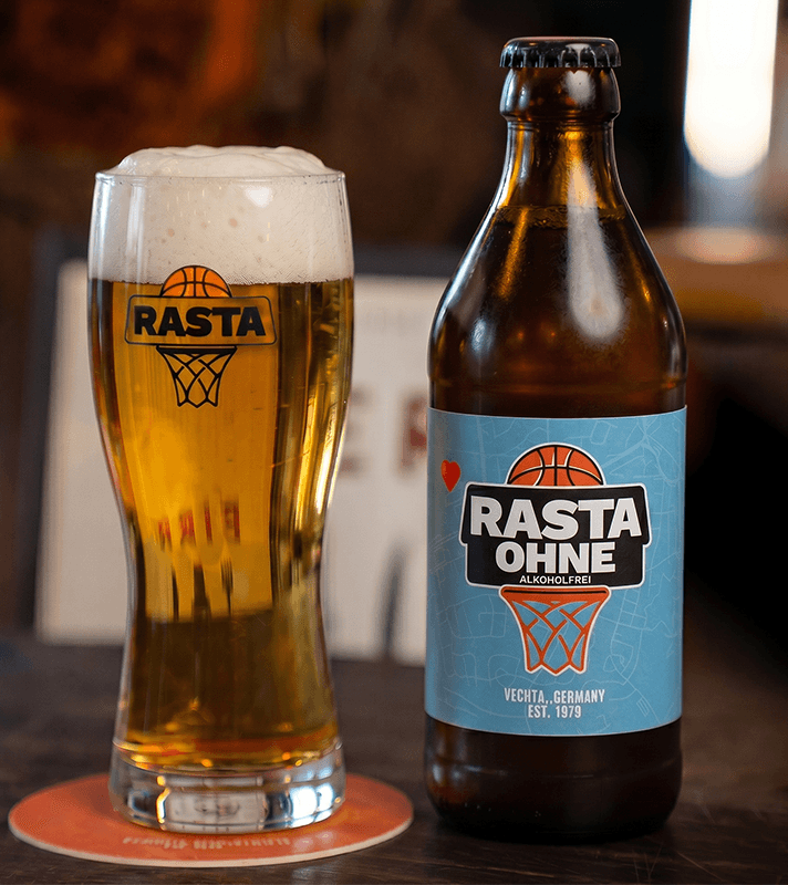 Eine braune Bierflasche mit Rasta Ohne-Logo in blau, daneben ein gefülltes Bierglas mit Schaumkrone und Rasta h-Logo, auf einem Holztablett.