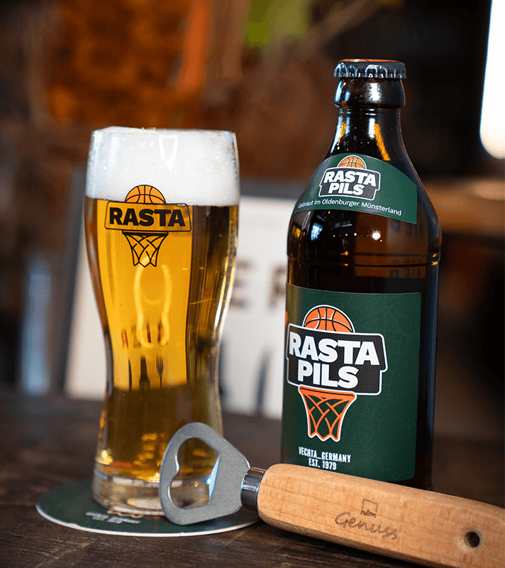 Bierflasche und Glas mit Rasta Pils, dazu ein Flaschenöffner und ein Untersetzer, alles auf einem Holztisch, im Hintergrund unscharfe Umgebung.