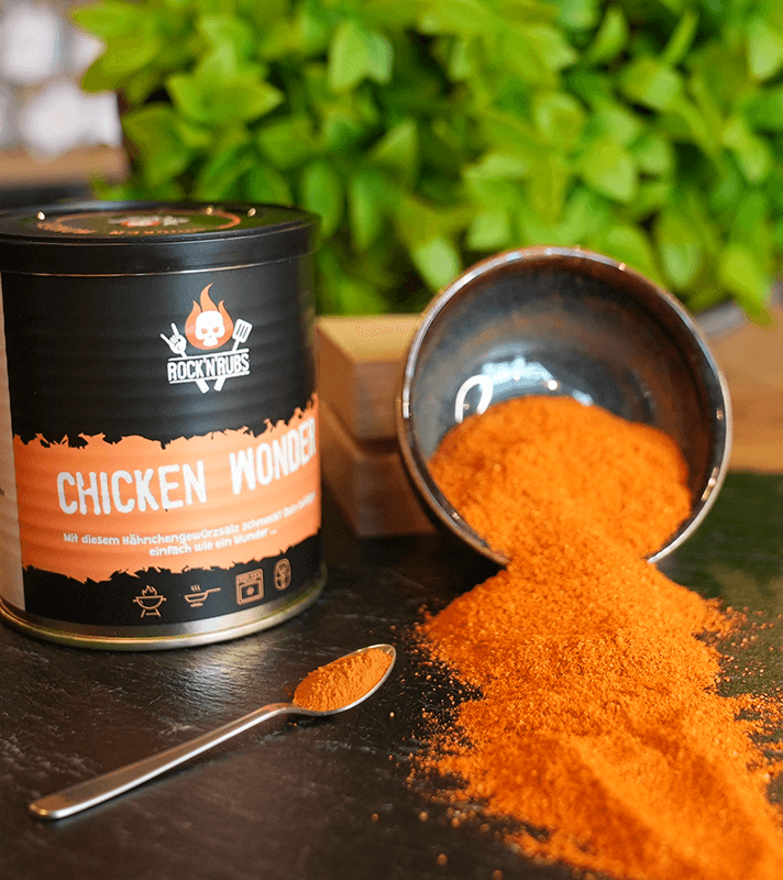 Hähnchen-Gewürzmischung "Chicken Wonder" von Rock N Rubs, orange Pulver in Schüssel und Dose, auf dunklem Holz, grüne Pflanzen im Hintergrund.