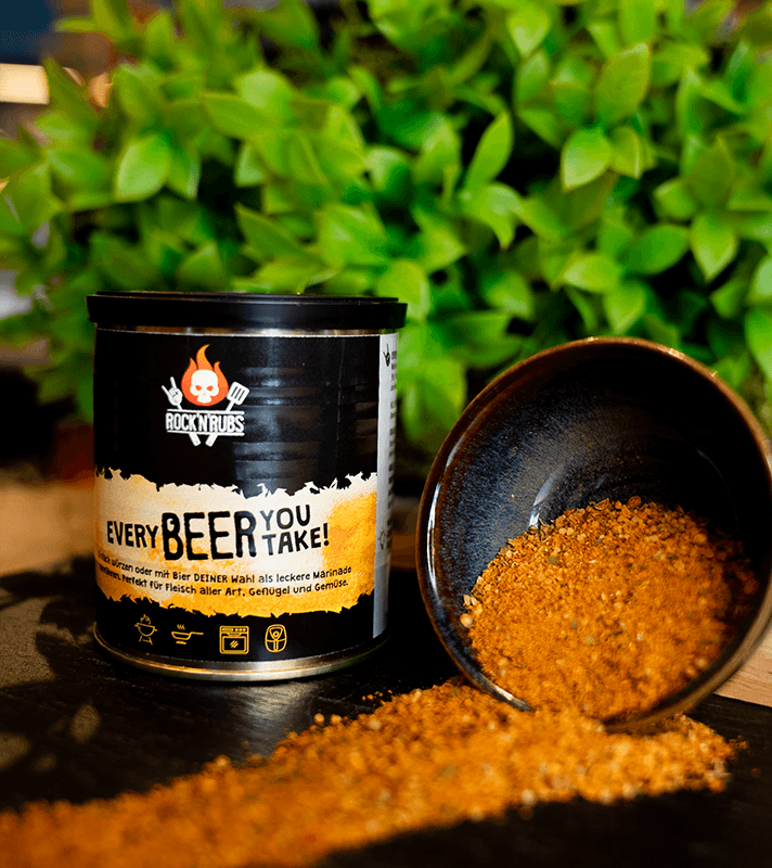Gewürzmischung in schwarzem Dosen mit "Rock N' Rubs" Logo, daneben eine Schale mit Gewürzpulver, auf dunklem Untergrund vor grünen Blättern.
