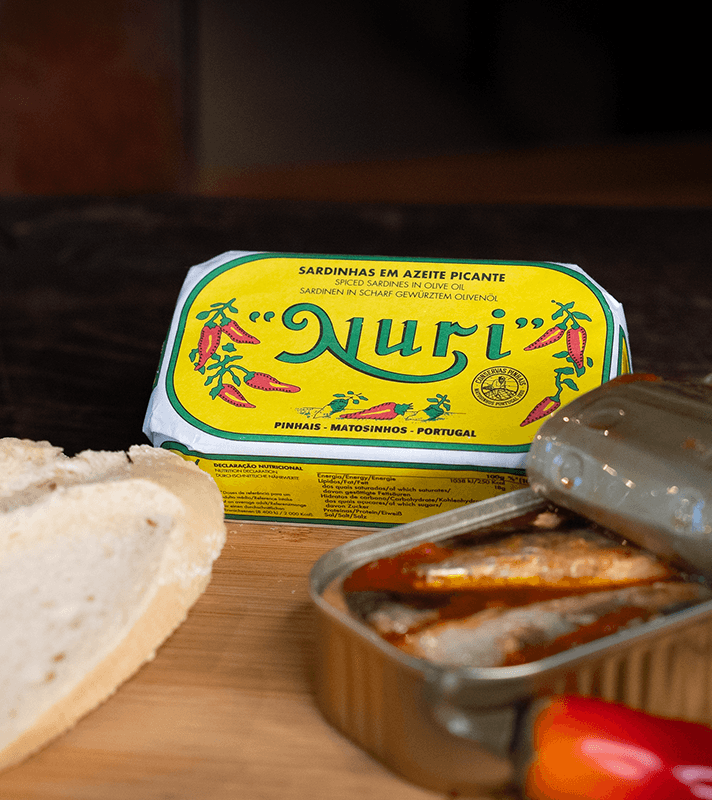 Dosen mit scharfen Sardinen in Olivenöl, eine Brotscheibe und eine geöffnete Dose mit Sardinen auf einem Holzbrett.