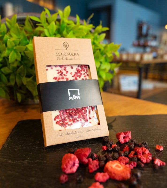 Weiße Schokolade mit Himbeerstückchen, verpackt in brauner Pappschachtel mit transparentem Fenster, auf schwarzem Steinplatte, im Hintergrund grüne Pflanze.