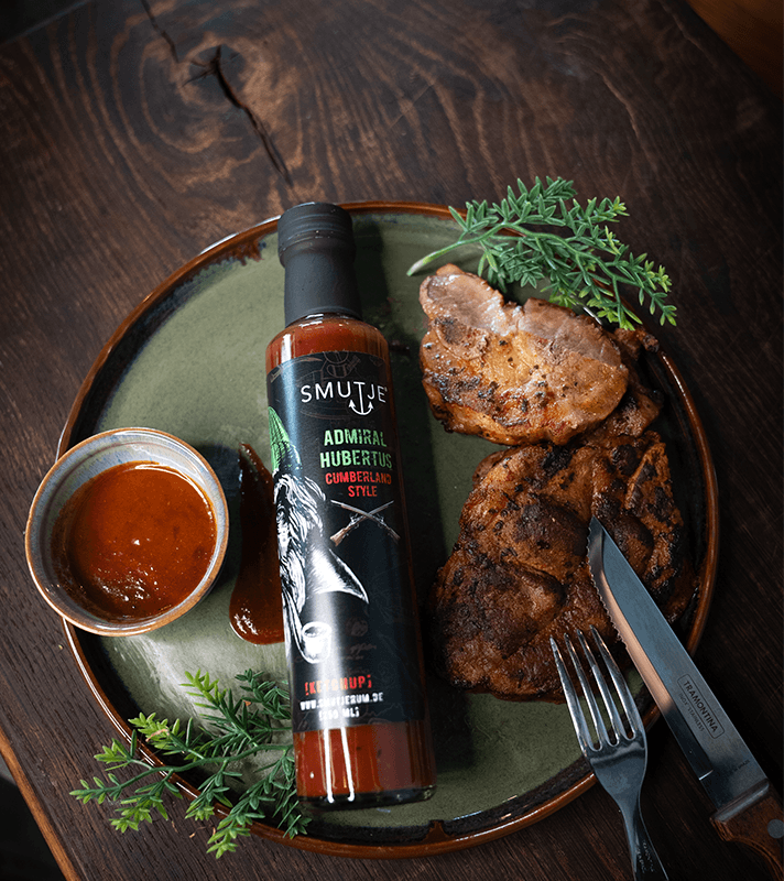 Gebratene Fleischstücke mit Grillmuster, grüne Kräuter, Flasche Smutje Admiral Huberts BBQ-Sauce, kleine Schüssel mit Sauce, dunkler Holzuntergrund.