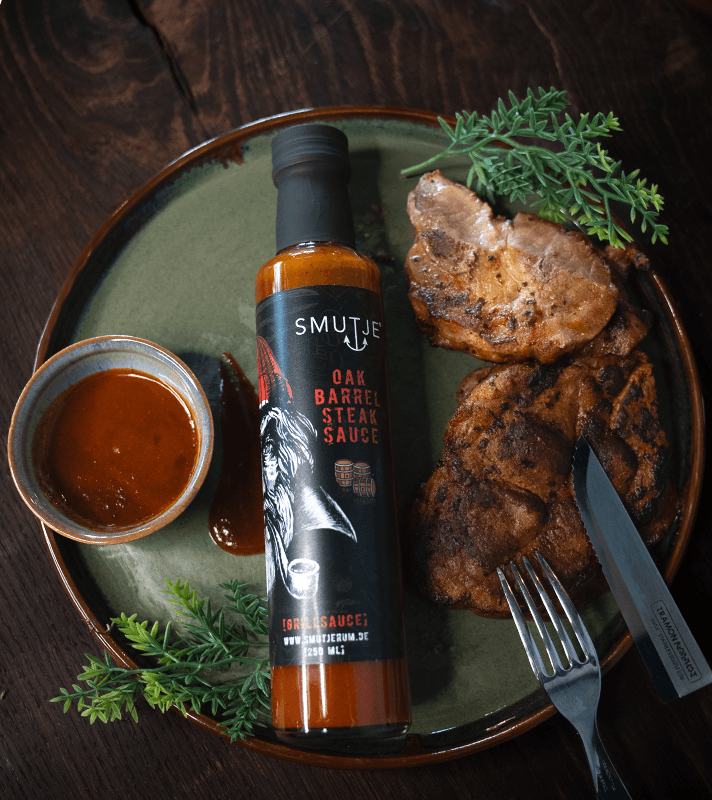 Alt-Text: Grillfleisch mit Knochen, grüne Kräuter, Flasche Smutje Oak Barrel Steak Sauce, kleine Schüssel mit Sauce, Messer und Gabel auf einem grünen Teller.