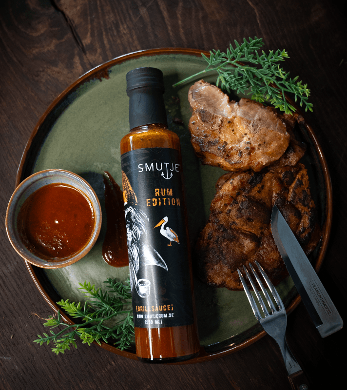 Brennendes Grillfleisch mit zwei gegrillten Schweinekoteletts, einer Flasche Rum-Edition Grillsoße, einer kleinen Schüssel BBQ-Sauce, grünen Kräutern und Besteck auf einem grünen Teller.