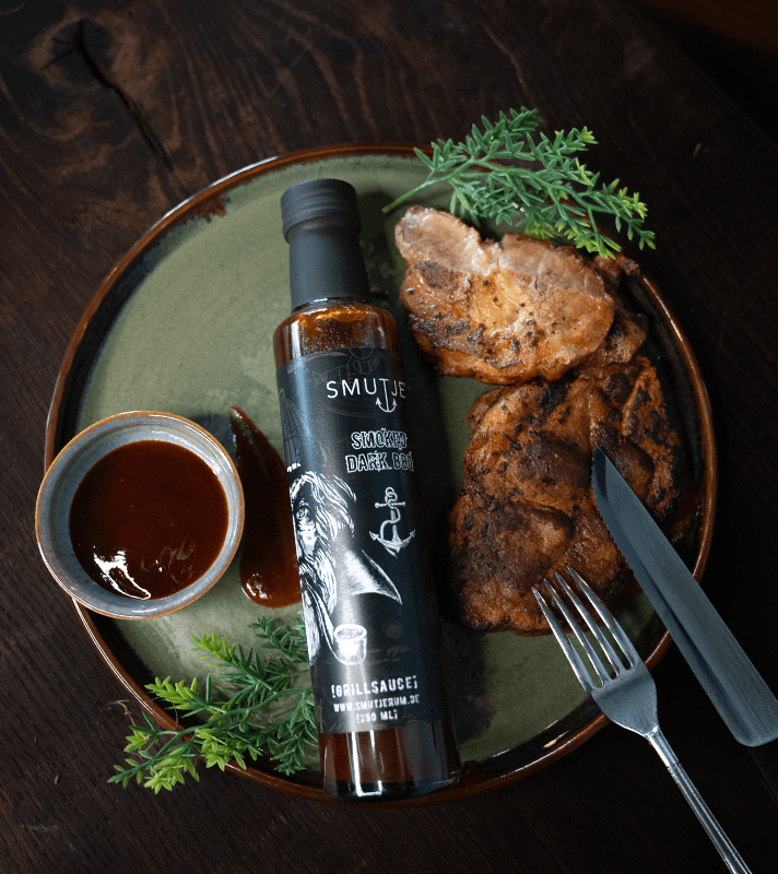 Gebratene Fleischstücke mit grünen Kräutern, dunkler BBQ-Sauce, einem Fläschchen Smutje Dark BBQ-Sauce, und einem kleinen Schälchen mit Sauce auf einem grünen Teller.