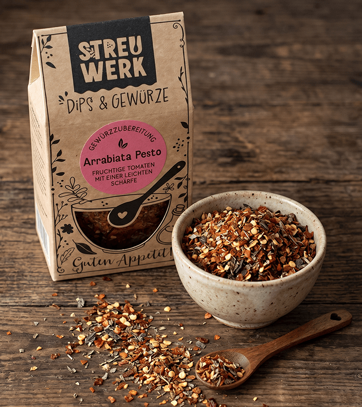 Kräuter- und Gewürzmischung in einer Schale und auf dem Holzlöffel, Verpackung mit "Streuwerk" und "Arrabiata Pesto" auf einem Holztisch.