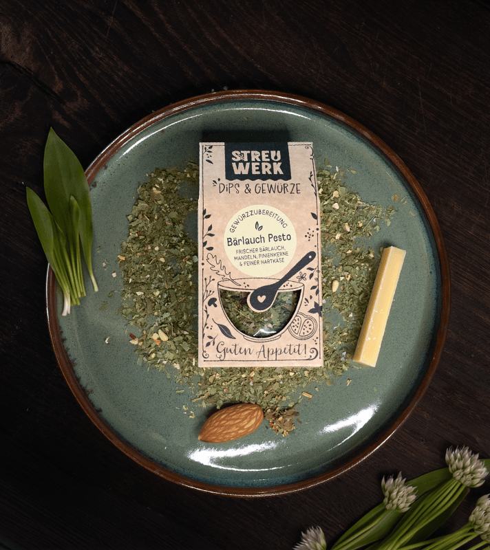 Getrocknete Kräutermischung mit Etikett, daneben eine weiße Schokolade, eine Mandel und frische Schnittlauch- und Gänseblümchen.