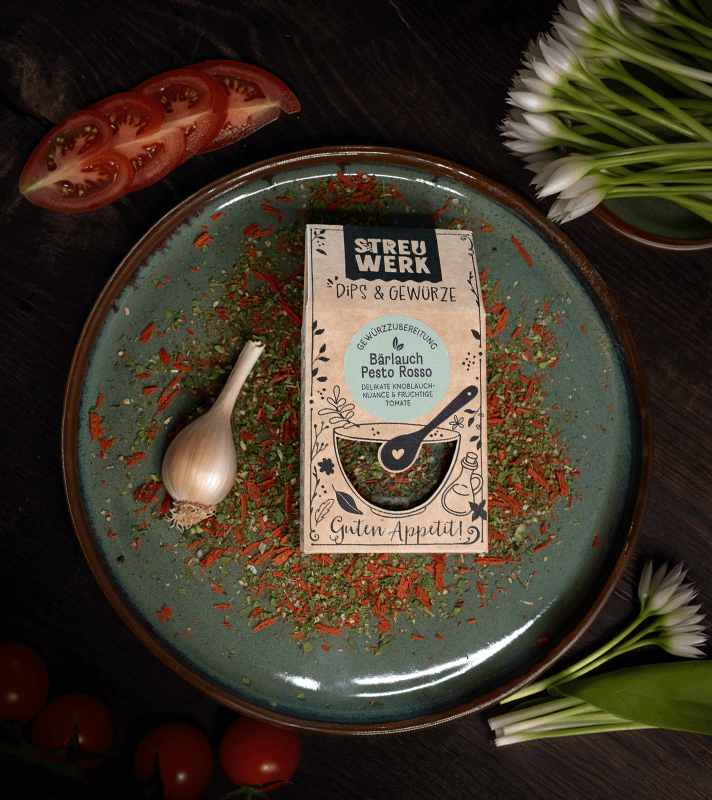 Bunte Gewürzmischung mit Knoblauchzehe auf einem blauen Teller, umgeben von Tomatenscheiben, weißen Blumen und dunklem Holz.