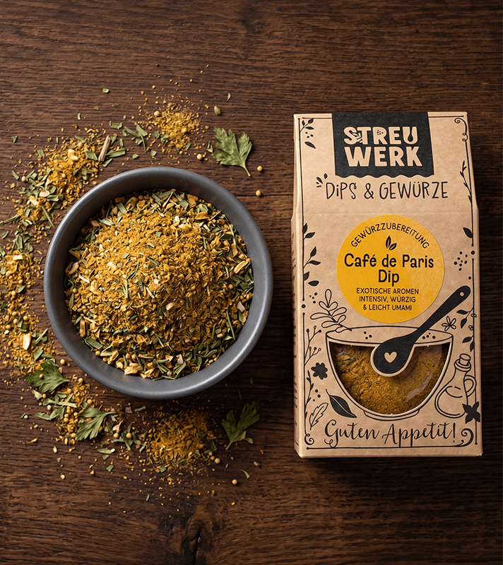 Paket mit Gewürzmischung "Café de Paris Dip" neben einer Schale mit gemahlenen Kräutern und Gewürzen, umgeben von Kräuterblättern und Gewürzresten auf einem Holzuntergrund.