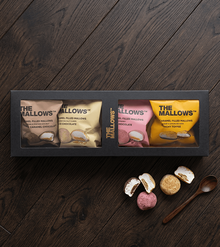 Schwarze Geschenkbox mit vier Sorten gefüllter Marshmallows in bunten Verpackungen, daneben drei Marshmallows, ein Holzlöffel auf dunklem Holztisch.