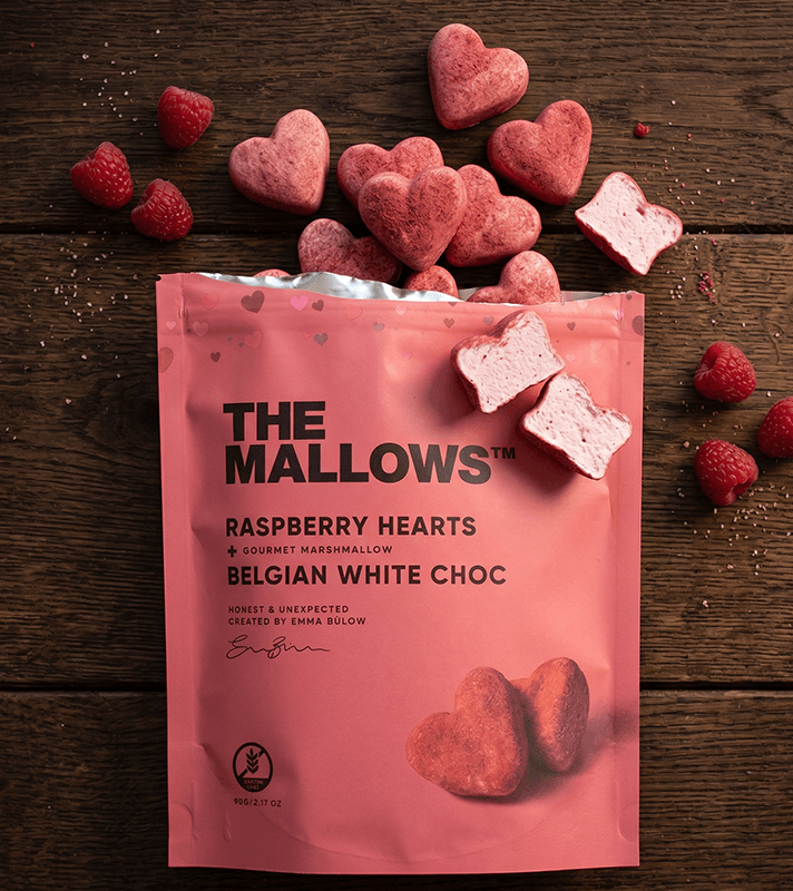 Raspberry hearts and marshmallows in pink packaging with chocolate, surrounded by raspberries and broken marshmallows on a wooden surface.