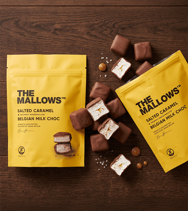 Zwei gelbe Verpackungen von "The Mallows" Salted Caramel belgischer Milchschokolade, umgeben von Schaumkuss-ähnlichen Süßigkeiten, Salzflocken und kleinen Karamellkugeln auf einem Holztablett.