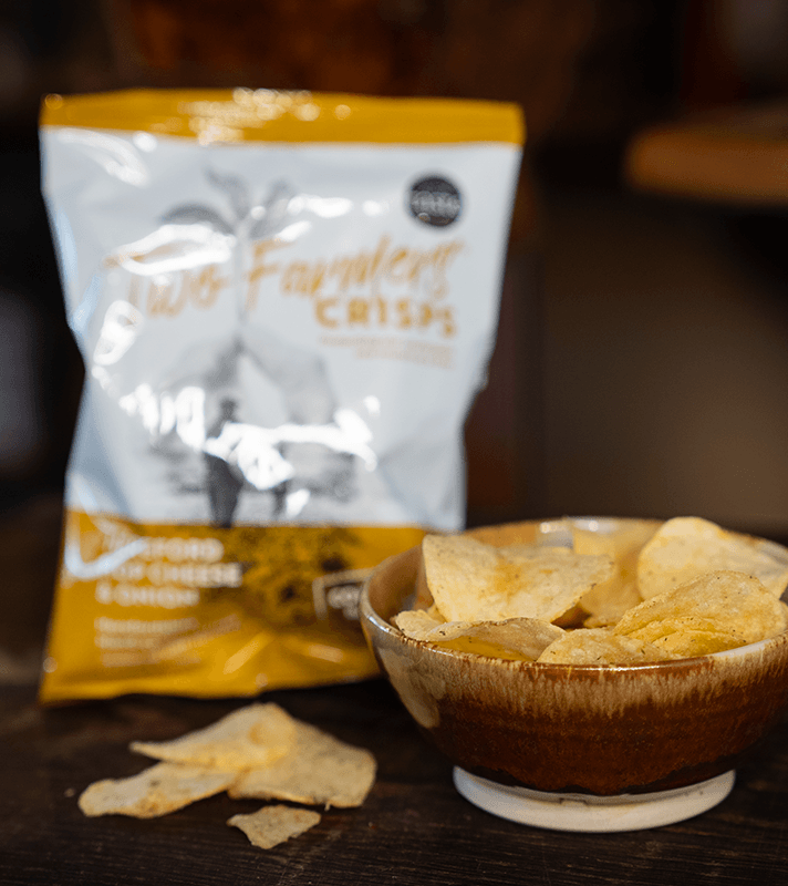 Knusprige Kartoffelchips in einer braunen Schüssel, im Hintergrund eine gelbe und weiße Packung mit "Farmers Crisps" aufgedruckt.