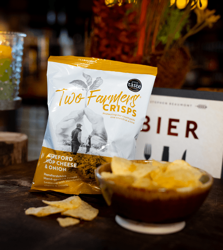 Knusprige Kartoffelchips in einer braunen Schüssel, im Hintergrund eine gelbe und weiße Packung mit "Farmers Crisps" aufgedruckt.