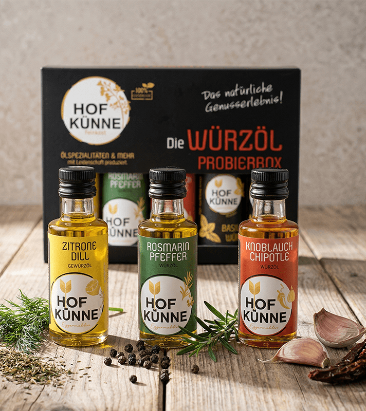 Drei verschiedene Würzöle umgeben von Knoblauch, Pfeffer und Rosmarin, im Hintergrund die Originalverpackung