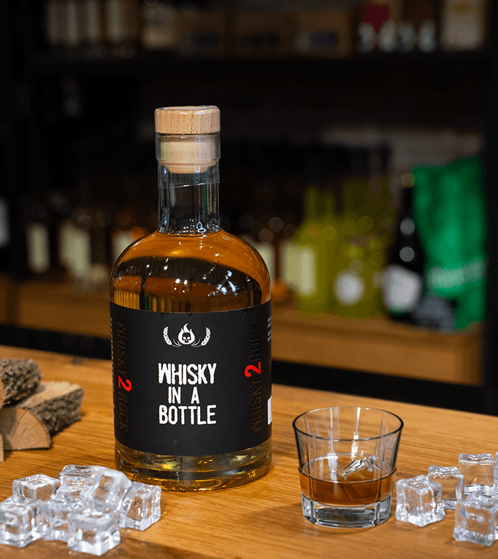 Flasche mit Whisky in einem Glas auf einem Holztablett, umgeben von Eiswürfeln, im Hintergrund eine Bar mit weiteren Flaschen.