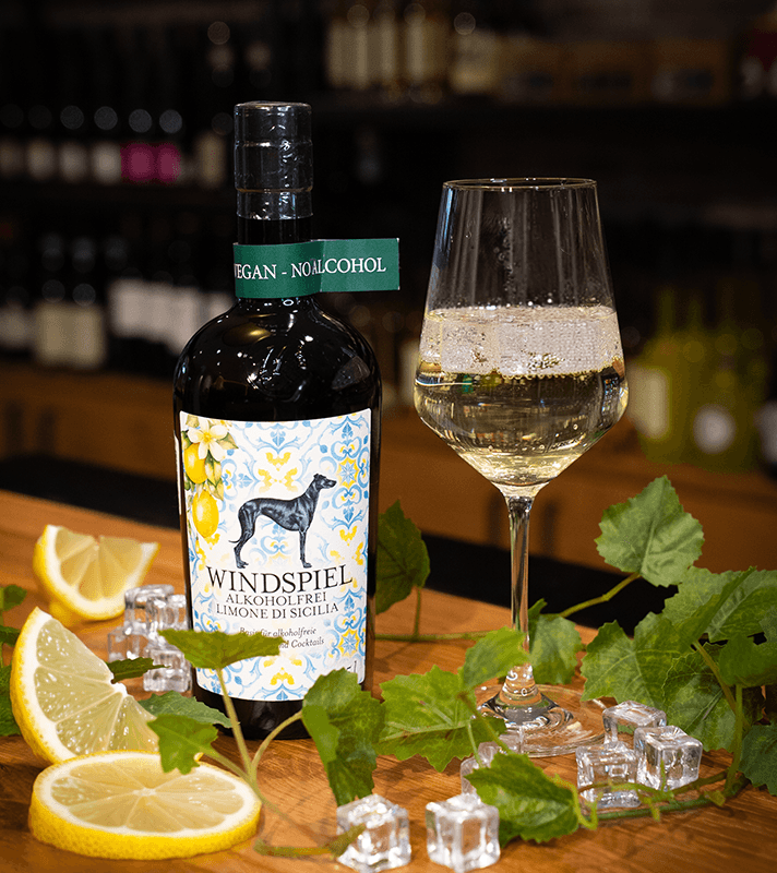 Alkoholfreier, veganer Zitronen-Limone-Drink in einem Weinglas, umgeben von Zitronenscheiben, Eiswürfeln und grünen Blättern auf einem Holztablett.